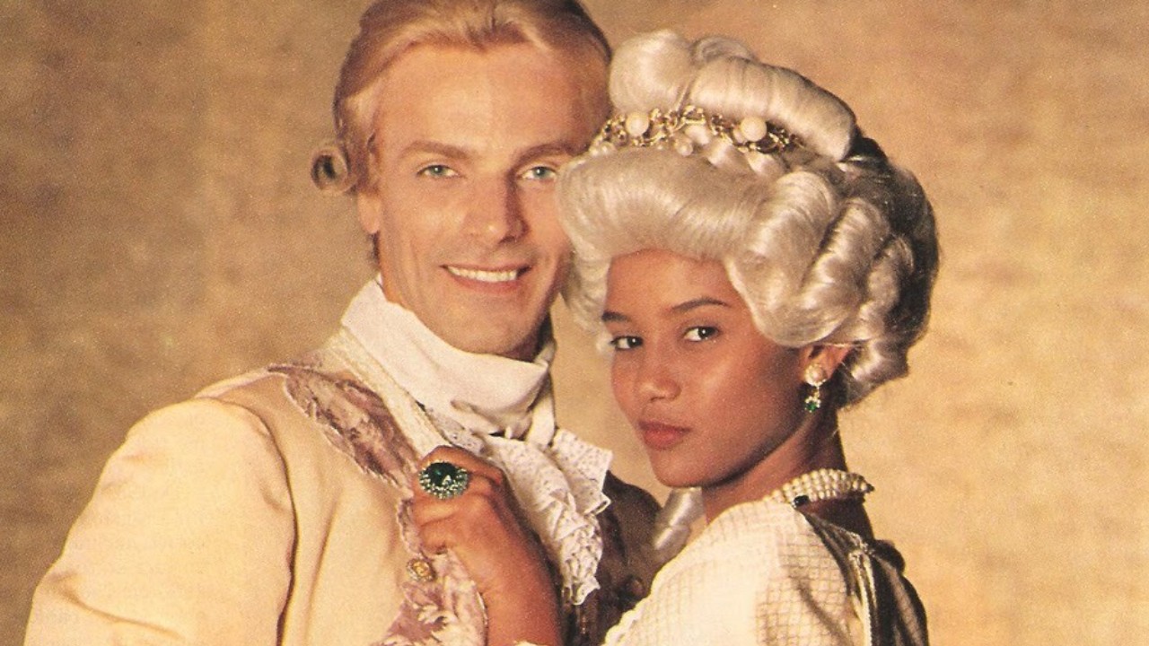 Taís Araujo como Xica e Victor Wagner como João Fernandes aparecem juntos, com roupas de época, perucas e acessórios característicos da novela Xica da Silva