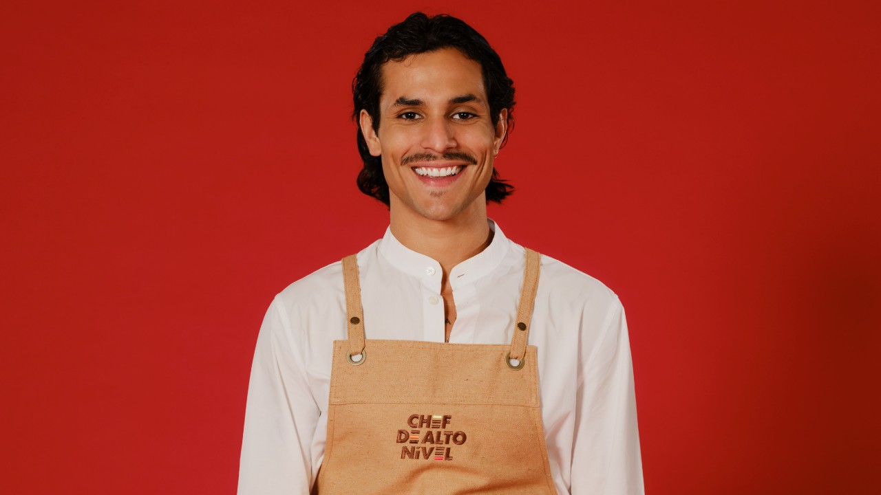 Yan Spadafori sorri com bigode, camisa branca e avental bege de Chef de Alto Nível, diante de um fundo vermelho