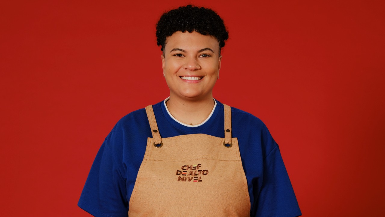 Yasmim Juliana sorri com cabelo curto, camiseta azul e avental bege de Chef de Alto Nível, diante de um fundo vermelho